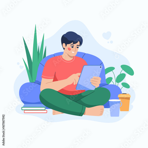 A young man sits comfortably cross-legged on a blue bean bag, using a digital tablet in a relaxed home environment with houseplants and a drink.