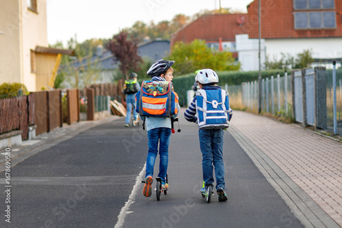 Two school kid boys in safety helmet riding with scooter in the city with backpack on sunny day. Happy children in colorful clothes biking on way to school.