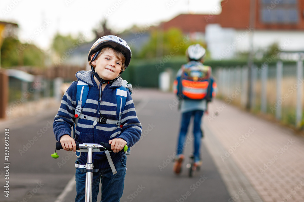 Obraz premium Two school kid boys in safety helmet riding with scooter in the city with backpack on sunny day. Happy children in colorful clothes biking on way to school.