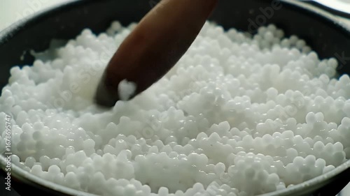 Detailed Close Up of Sago Pearls Being Roasted in Pan with Wooden Spoon Stirring Action Showcasing Translucent White Grains Perfect For Cooking Process and Culinary Themes and Recipe Illustrations