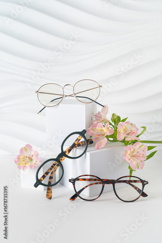 Three pairs of plastic eyeglass frames on white background with flowers. Eyewear fashion collection. Trendy eyeglasses still life minimal style. Optic store discount sale, promotion. Vertical
