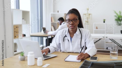 Wallpaper Mural Confident young woman doctor using laptop and writing prescription in modern medical office. Bright workspace featuring healthcare equipment, teamwork depicted with colleague in background Torontodigital.ca