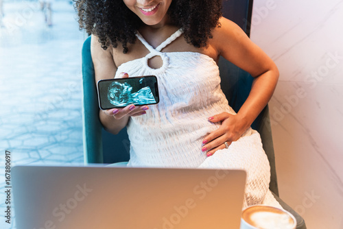 Anonymous smiling pregnant woman showing baby ultrasound picture to laptop