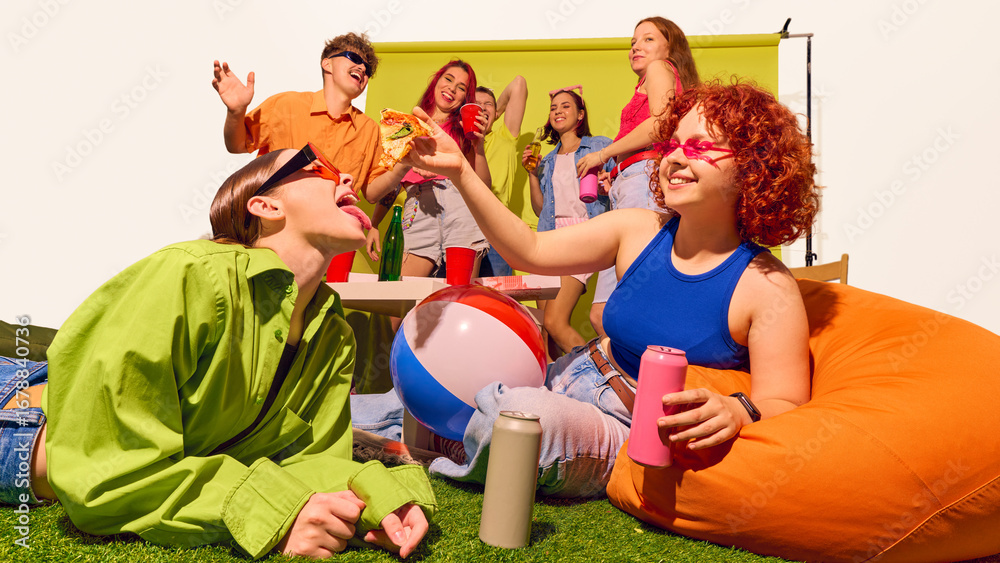 Obraz premium Young woman feeding pizza to friend on grass with group cheering behind. Concept of playful intimacy, carefree lifestyle, gen z friendship, vibrant pop culture, and joy in togetherness.