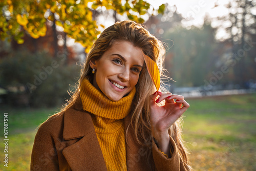 Beautiful charismatic  blonde woman holding yellow leaf in a park, wearing turtleneck sweater and smiling, autumn season portrait at sunset 