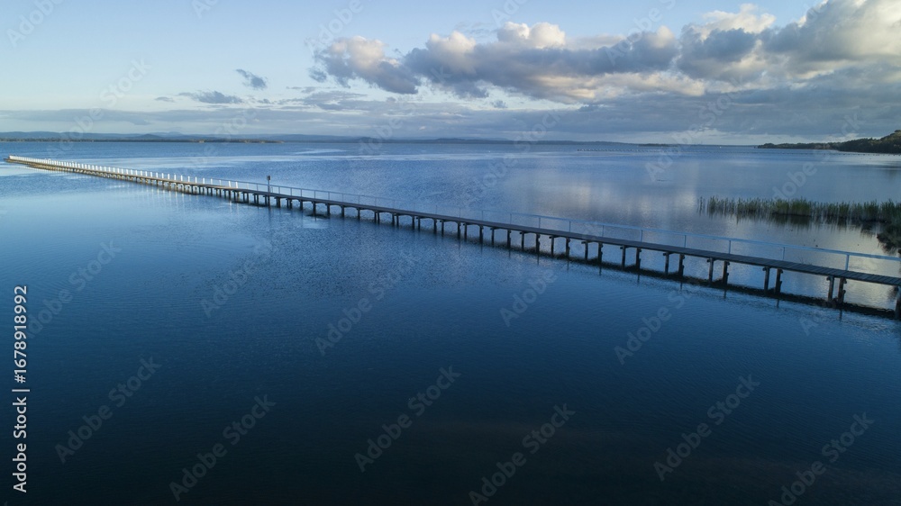 Aerial view of the long jetty stretching far into the shimmering, dark blue waters under a serene sky, Lakes Entrance, Victoria, Australia. Aerial view of the long jetty stretching far into the shimmering, dark blue waters under a serene sky, Lakes Entrance, Victoria, Australia.