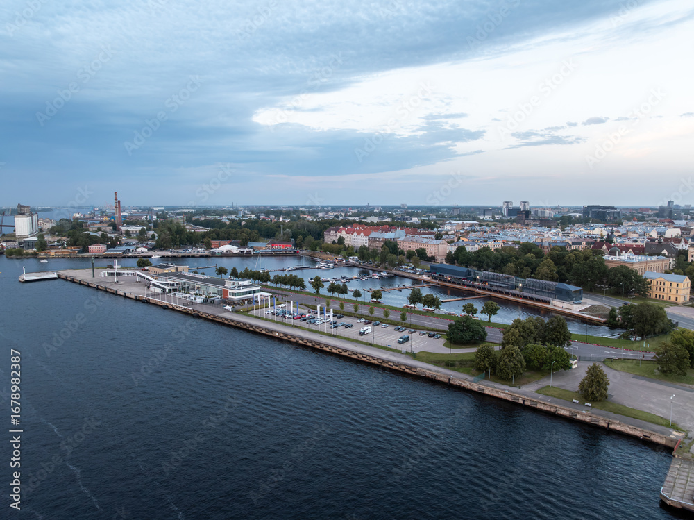 Obraz premium Tallinn waterfront with a marina, docked boats, a paved promenade, parking spaces, colorful buildings, a tall chimney, and a cloudy sky.
