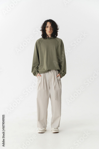 Man in green sweatshirt and beige pants standing in minimalistic studio setting.