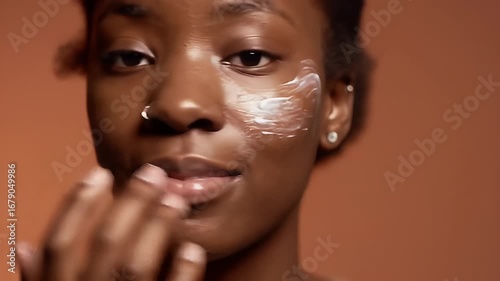 Serene Young African American Woman Applying Cream to Her Face in Close Up with a Natural Expression Against a Warm Brown Background for Beauty and Skincare Product Advertisement and Promotion