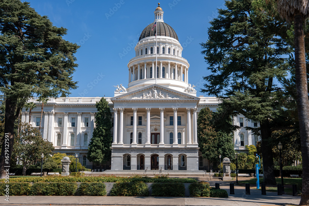 Obraz premium The California State Capitol in Sacramento features a white neoclassical facade, tall columns, a golden topped dome, and surrounding greenery under a clear sky.