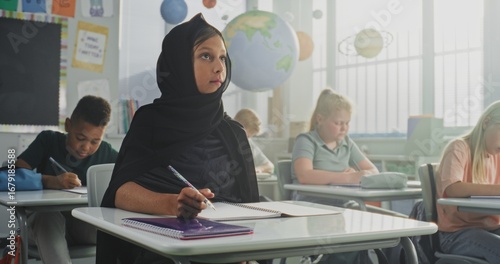 Canvas Print Muslim Primary School Girl Wearing Hijab Sitting at the Desk, Doing Task in Notebook During Lesson or Lecture, Writing School Test