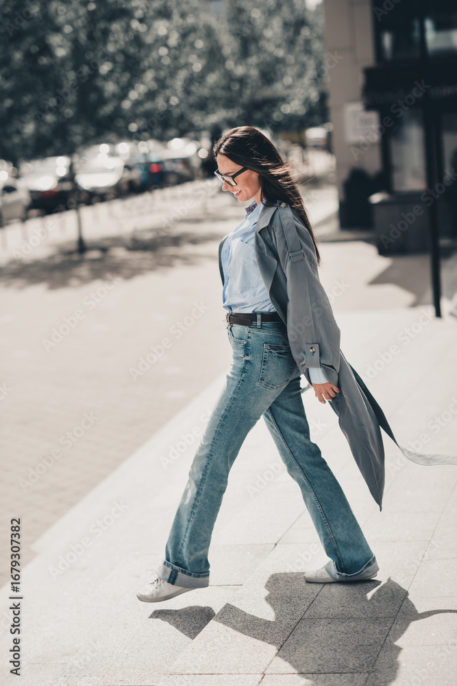 Fototapeta premium Stylish brunette woman in a grey trench coat walking confidently in an urban street setting under sunlight outdoors, vertical portrait
