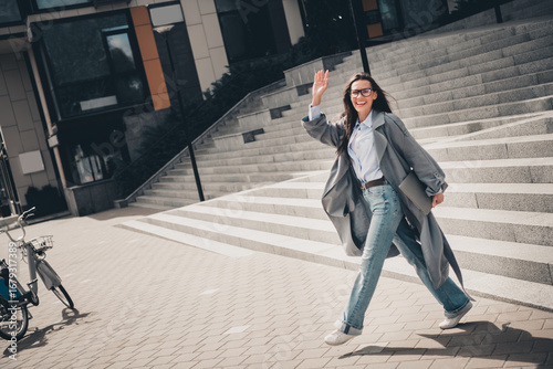 Stunning woman in urban outdoors with a charming smile, wearing a stylish grey trench coat and jeans on a sunny day.