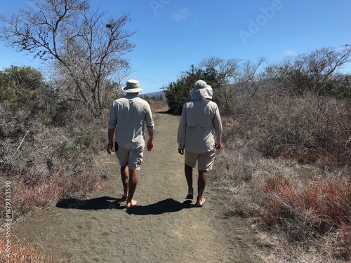 Two barefoot Galapagos tour guides in beige uniforms walking down a sandy track through dry island vegetation, back view.