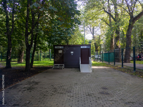 Small modular restroom in a city park with signage and an accessibility ramp by a paved brick path