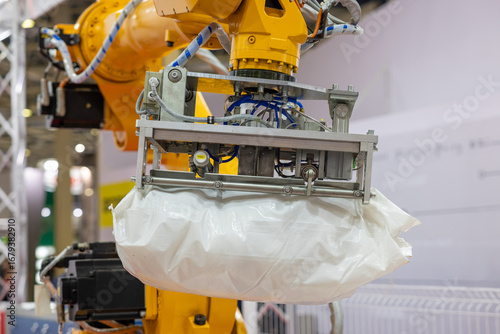 An industrial robotic arm with a pneumatic gripper lifts a heavy white bag in a factory setting. The background shows a manufacturing environment