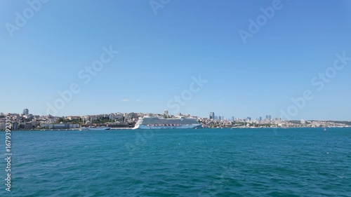 A large cruise ship glides smoothly across turquoise waters next to a lively city skyline. Buildings rise alongside the shore against a bright sunny backdrop.