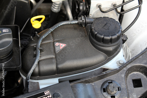 Close-up black plastic car coolant reservoir with cap and warning symbols, located in engine bay. Container has matte texture and connects to surrounding components via hoses. Сar cooling system.