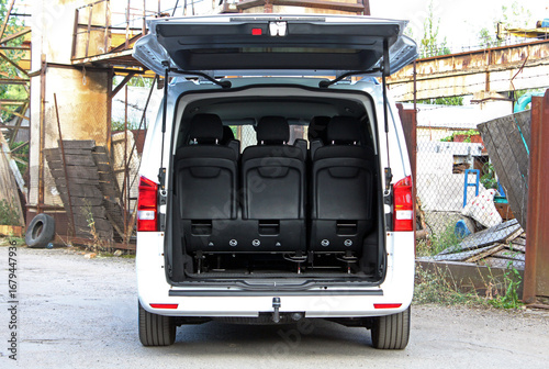 Mini van open trunk. Passenger van trunk. Rear view of white minivan with trunk door open, showing black passenger seats in upright position. Vehicle is parked outdoors near rusty industrial structure