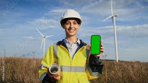 Beautiful Caucasian woman with head protection and high-visibility vest standing in front of rotating wind turbine. Showing to camera smartphone with green screen. Chroma key. Smiling positively.
