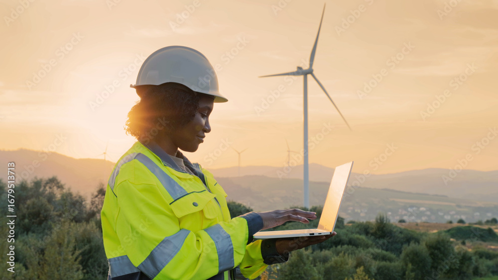 custom made wallpaper toronto digitalSide view of African American woman working at wind power station. Wearing yellow protective jacket with gray-green inserts and safety helmet. Using laptop for productive work. Renewable green energy.