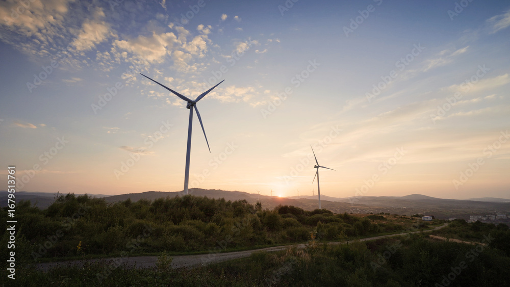 Obraz premium Beautiful view of two wind turbines slowly rotating on top of hill. Blue sky. Bright sunset visible on horizon. Green energy production concept. Caring about nature. Healthy planet.