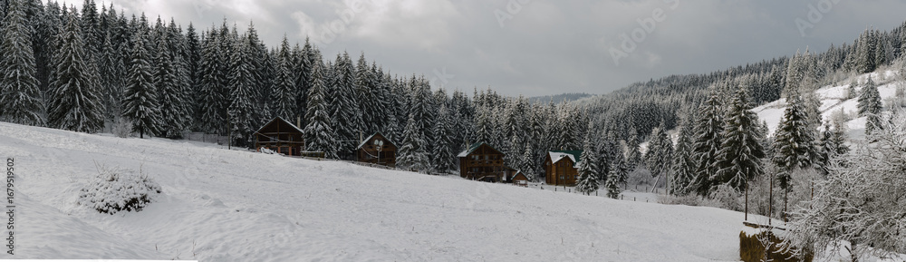 Naklejka premium Small wooden chalets peek through the snow-covered trees, evoking warmth and coziness. The cloudy sky adds a soft, tranquil light, emphasizing the fairy-tale charm of the winter
