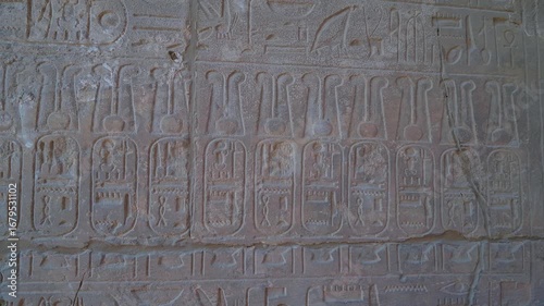 Close up footage showcasing intricate ancient Egyptian hieroglyphs carved into a stone wall, likely within a temple. The detailed relief carvings depict symbols, figures, and texts from an ancient civ