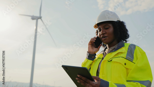 Focused African American woman talking with someone on smartphone while looking at tablet device. Remote communication concept. Power plant worker in protective equipment completing maintenance check.