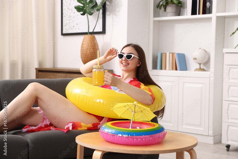 Obraz premium Happy woman in sunglasses with mason jar of juice and inflatable ring on sofa at home. Dreaming about vacation at sea