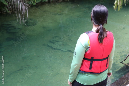 
A woman observing a spring of crystal clear water with greenish tones, being able to see the rocks to the bottom of the lake and around coconut trees and a bridge.