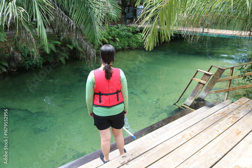 
A woman observing a spring of crystal clear water with greenish tones, being able to see the rocks to the bottom of the lake and around coconut trees and a bridge.