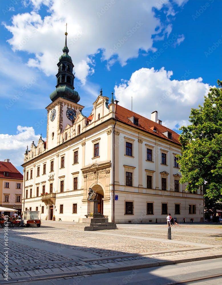 Fototapeta premium A magnificent, light-beige historical building, showcasing a tall, dark-green turret, stands proudly in a sun-drenched square, with a statue at its base, and mature trees casting a gentle shade.