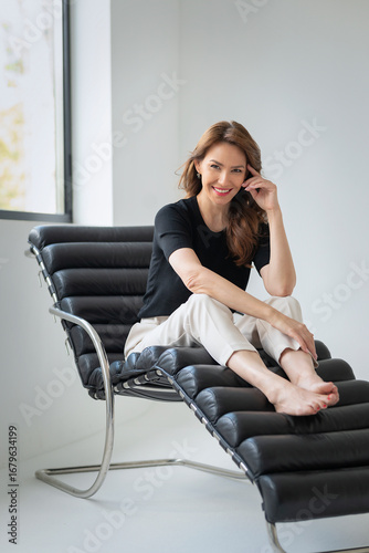 Full length of a brunette haired woman sitting in a modern chair in an empty white room