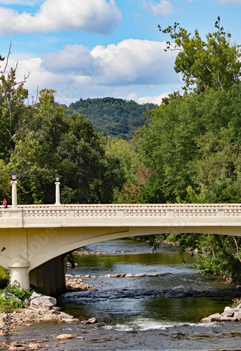 Looking down a stream as water flows under an arched white bridge with a blue sky and fluffy clouds