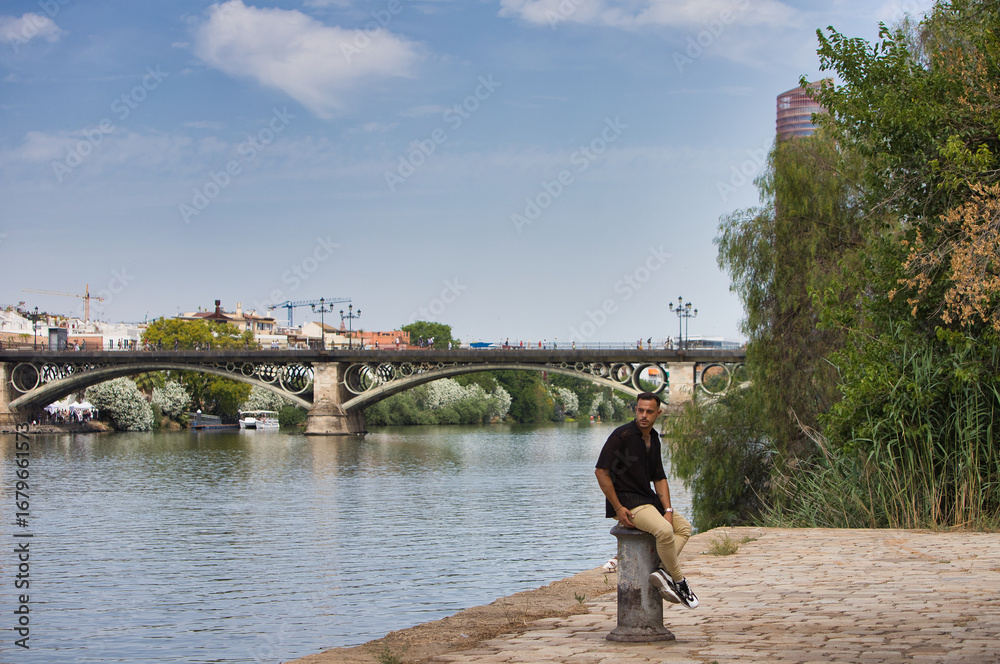 Fototapeta premium Young, handsome man with a beard, wearing a black shirt and beige cargo trousers, sitting on a metal poster used to moor boats,with the river and the famous Triana Bridge in Seville in the background.