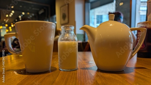 A white tea service comprising of a mug, tea pot and a mini milk bottle on a varnished wood table in a large dining area. 