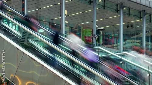 People ascend and descend an escalator with a long exposure motion blur effect, creating streaks of color and movement. This modern urban environment features glass panels, reflective surfaces, and