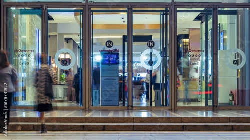 Blurred figures of people enter and exit a modern building through automatic glass doors, creating a sense of dynamic motion in a busy urban environment. The scene captures the constant flow of