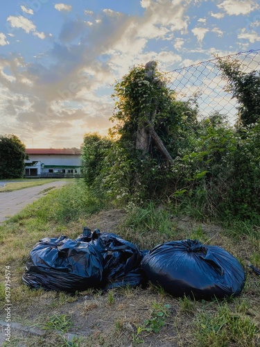 Two black garbage bags lie on overgrown grass beside a rural fence and sidewalk, captured during golden hour. Symbolic image of littering, waste management, and environmental impact in suburban areas.