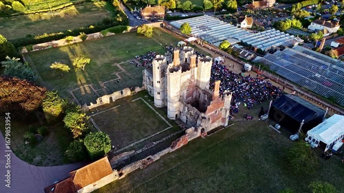 aerial drone shot mid angle flying over towards ancient old English Titchfield abbey ruins with large crowd of people in countryside in summer at sunset