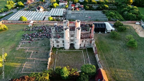 aerial high wide angle shot face on flying over panning down on ancient old English Titchfield abbey ruins in countryside with large crowd of people in summer at sunset