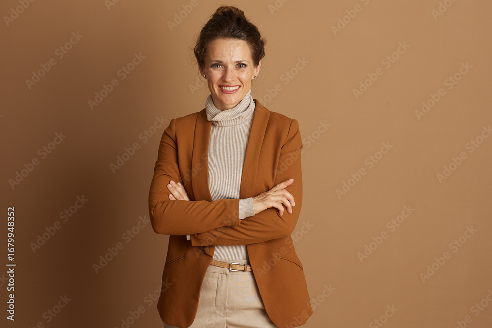Obraz premium A confident businesswoman with bun hairstyle smiles at the camera, arms crossed. Wearing brown blazer and beige turtleneck, she exudes professionalism and approachability against warm brown background