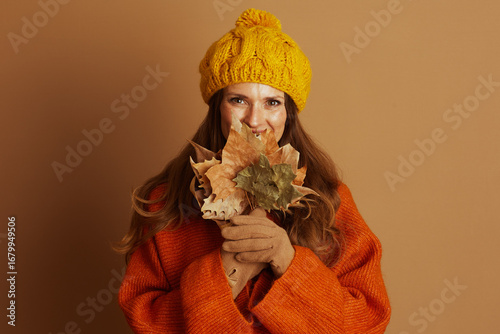 A middle-aged woman in a yellow beanie and orange sweater smiles brightly, holding autumn leaves to her lower face while gazing at the camera. This cozy portrait exudes cheerfulness.