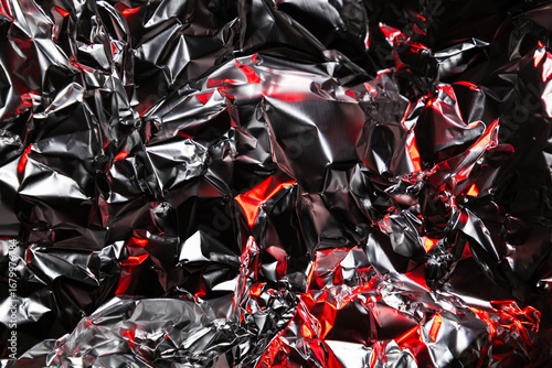 Close up of crumpled aluminum foil with shiny silver surface and dramatic red highlights. Abstract metallic background with reflective texture and artistic light play.