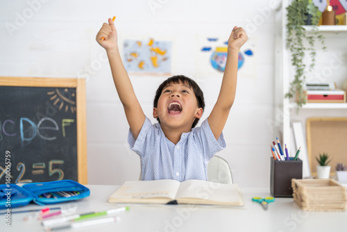 Excited young asian boy celebrating success while studying at home, raising hands in joy with open notebook, creative classroom, education and happy school moment, celebrating academic achievement