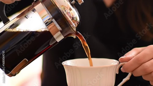 A person pours freshly brewed coffee from a French press into a white teacup.