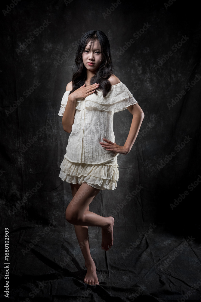 Fototapeta premium Young Asian woman standing and smiling in studio, wearing off-shoulder white blouse and layered skirt, posing gracefully with one hand on chest and one leg raised, dark background.