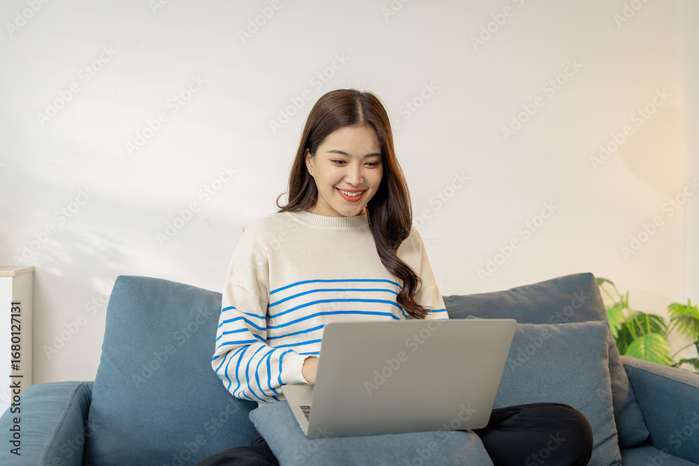 Naklejka premium A young woman is sitting comfortably on a sofa using a laptop, smiling with focus and joy. She looks engaged in her work or online communication, creating a warm lifestyle concept.