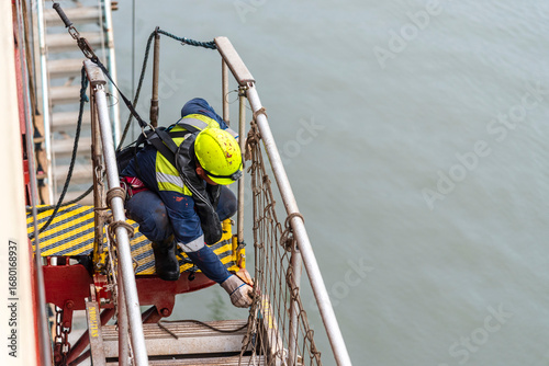 A deckhand prepares the pilot ladder as the ship readies for port entry. Secured in a safety harness, he carefully ensures the setup is ready, focusing on the task in a high-risk environment. 